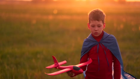 The-boy-in-the-costume-of-a-super-hero-running-in-a-red-cloak-laughing-at-sunset-in-summer-field-representing-that-he-was-the-pilot-of-the-plane